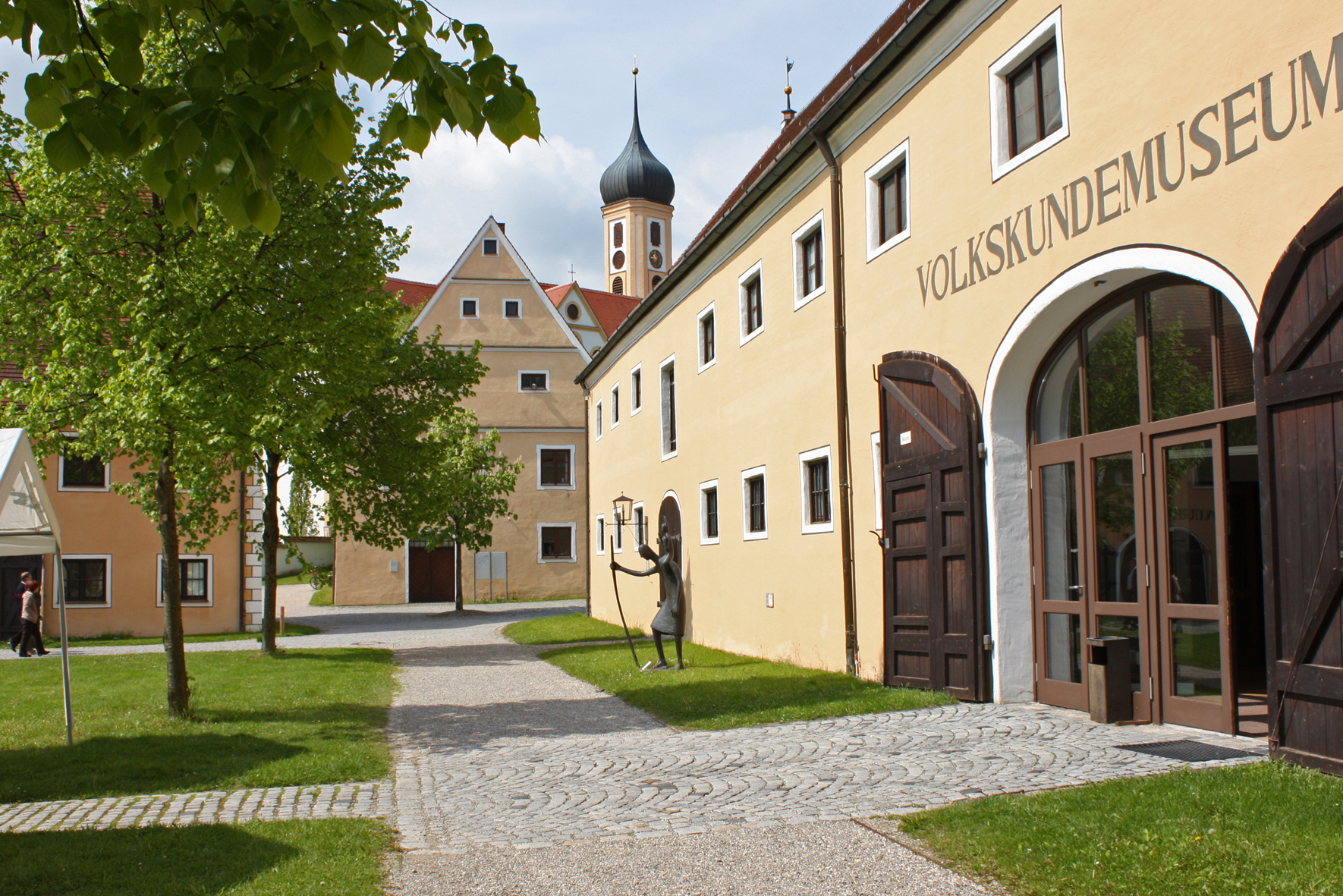 Kirchturm und mehrere ockerfarbene Museumsgebäude und Weg mit Wiese und Bäumen
