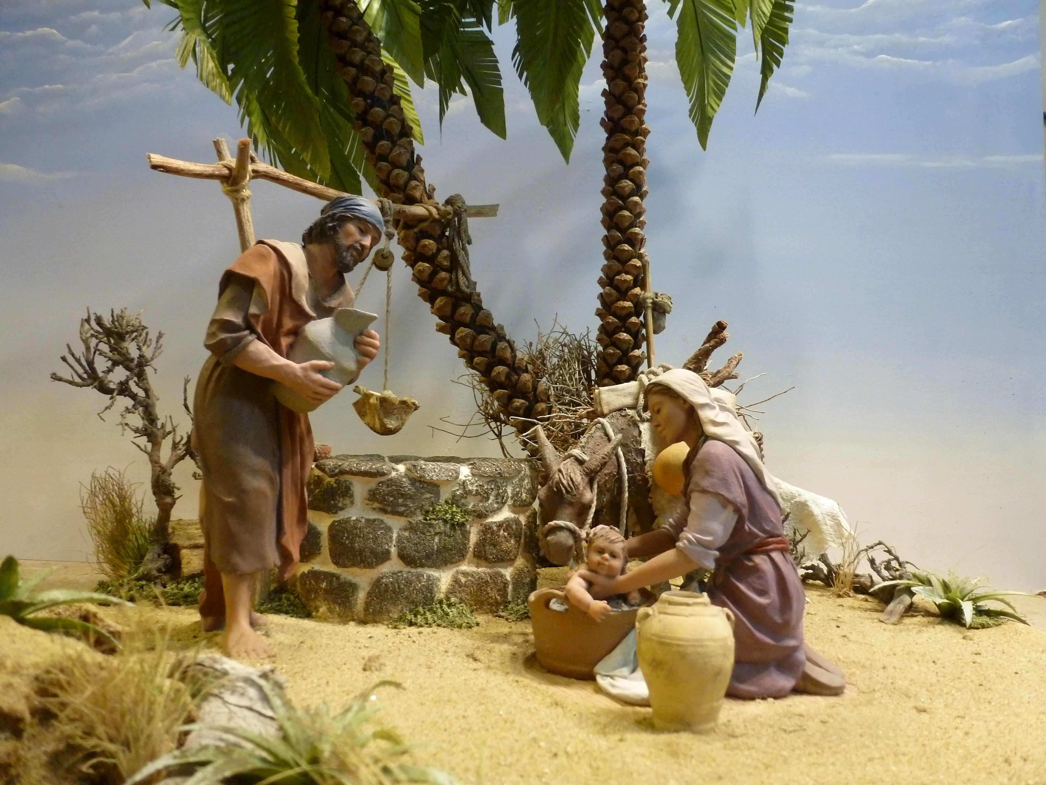 mit steinen umrandeter Brunnen, dahinter zwei Palmen, davor Maria, die das Jesuskind badet und Josef, der Wasser im Krug berbei bringt