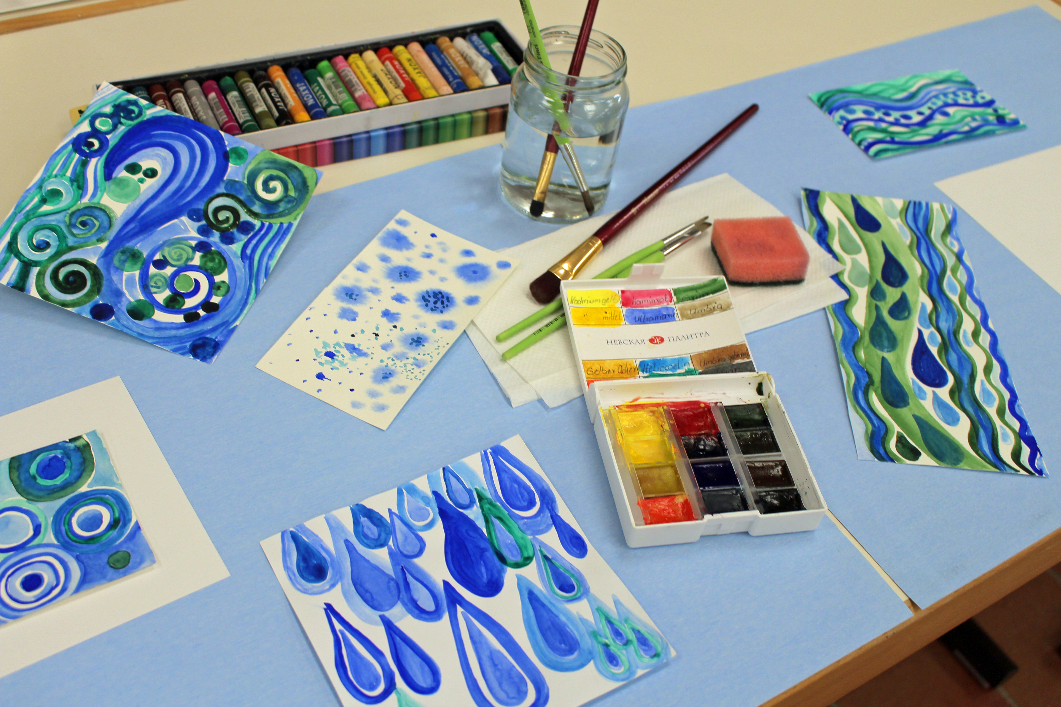 in Blau- und Grüntönen bemalte Bilder neben Pinsel, Wasserglas und Malfarben