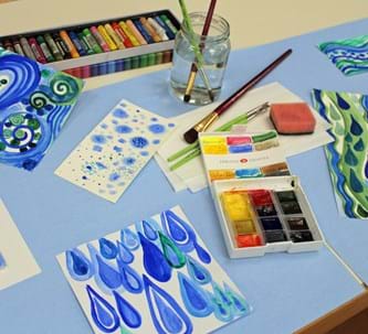 in Blau- und Grüntönen bemalte Bilder neben Pinsel, Wasserglas und Malfarben